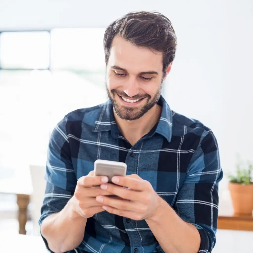 A smiling man with a beard wearing a blue checked shirt sits indoors, looking down at his mobile phone after LASIK surgery. A small potted plant and blurred background are visible behind him.