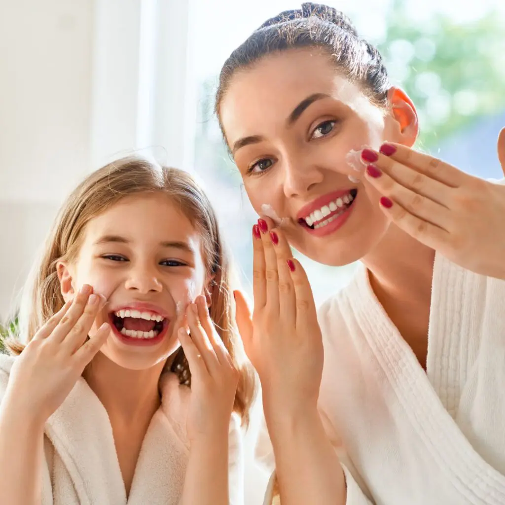 A smiling woman and young girl wearing white bathrobes apply cream to their faces together in a bright bathroom, enjoying a skincare routine.