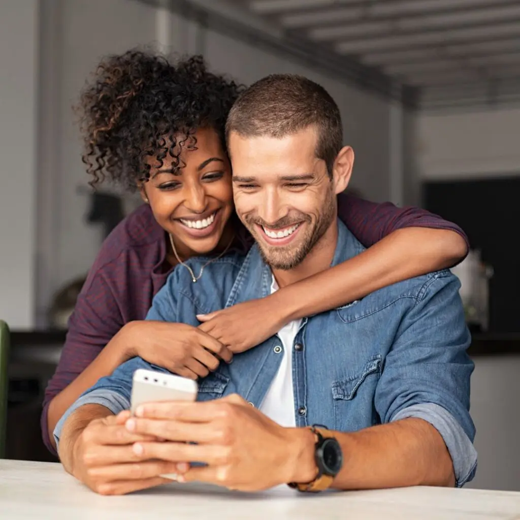 A smiling couple sit at a table, with the woman hugging the man from behind as they both look happily at a mobile phone in his hands.