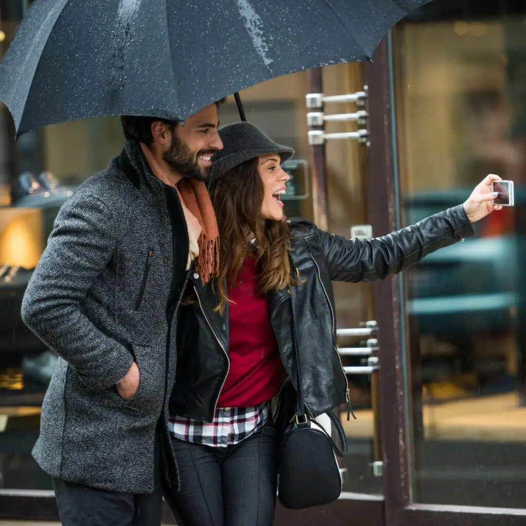 A smiling couple stands under a black umbrella, taking a selfie together on a rainy day. Both wear coats and hats, and appear happy as they pose outside a shop.