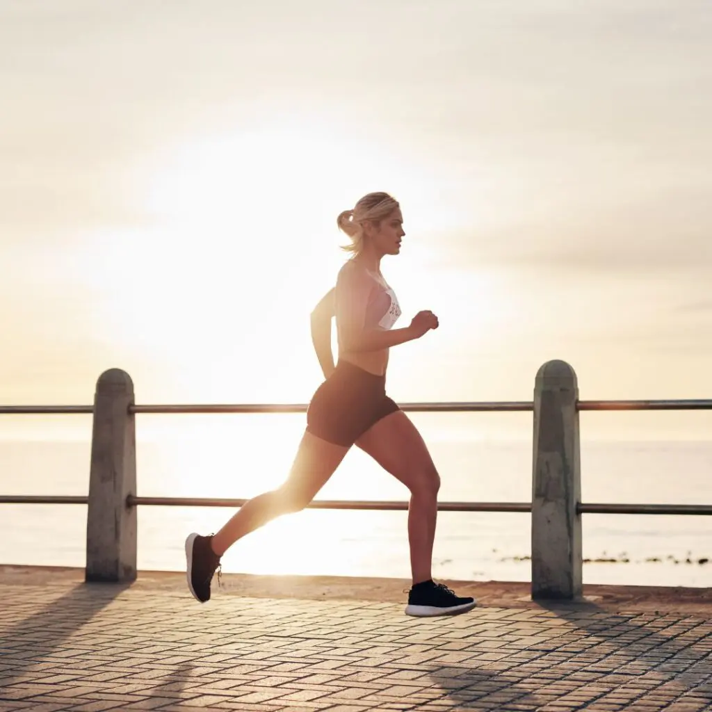 A woman jogs along a paved path by the sea at sunrise or sunset, with the sun low in the sky behind her and railings separating the path from the water.