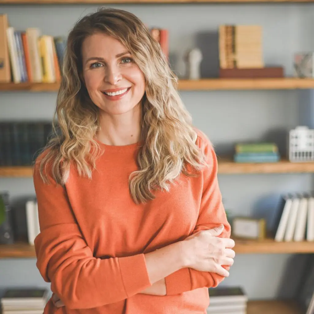 A woman with wavy blonde hair, wearing an orange jumper, smiles whilst standing in front of a book-filled wooden shelf in a well-lit room.
