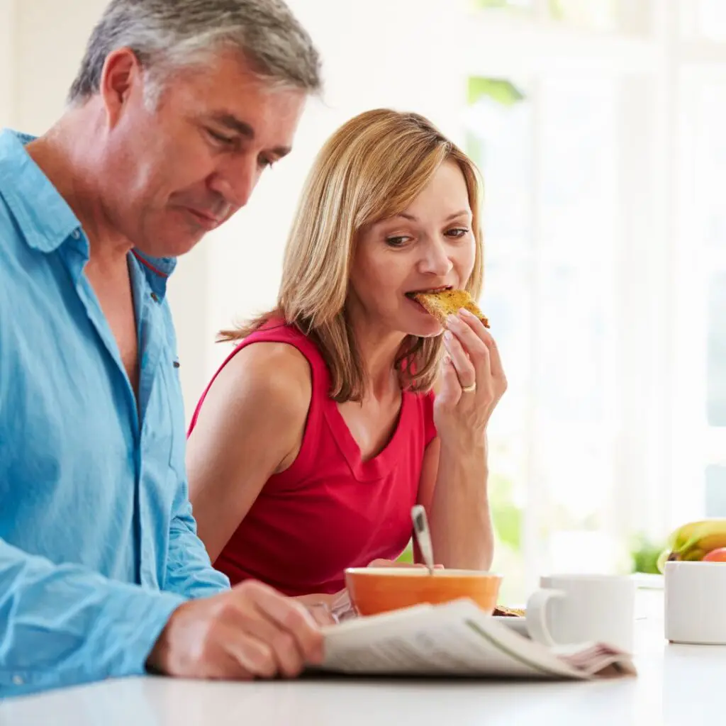 A middle-aged man reads a newspaper while a woman next to him eats toast and smiles. They are sitting at a white breakfast table with a bowl, mug, and fruit in a bright, sunlit kitchen.