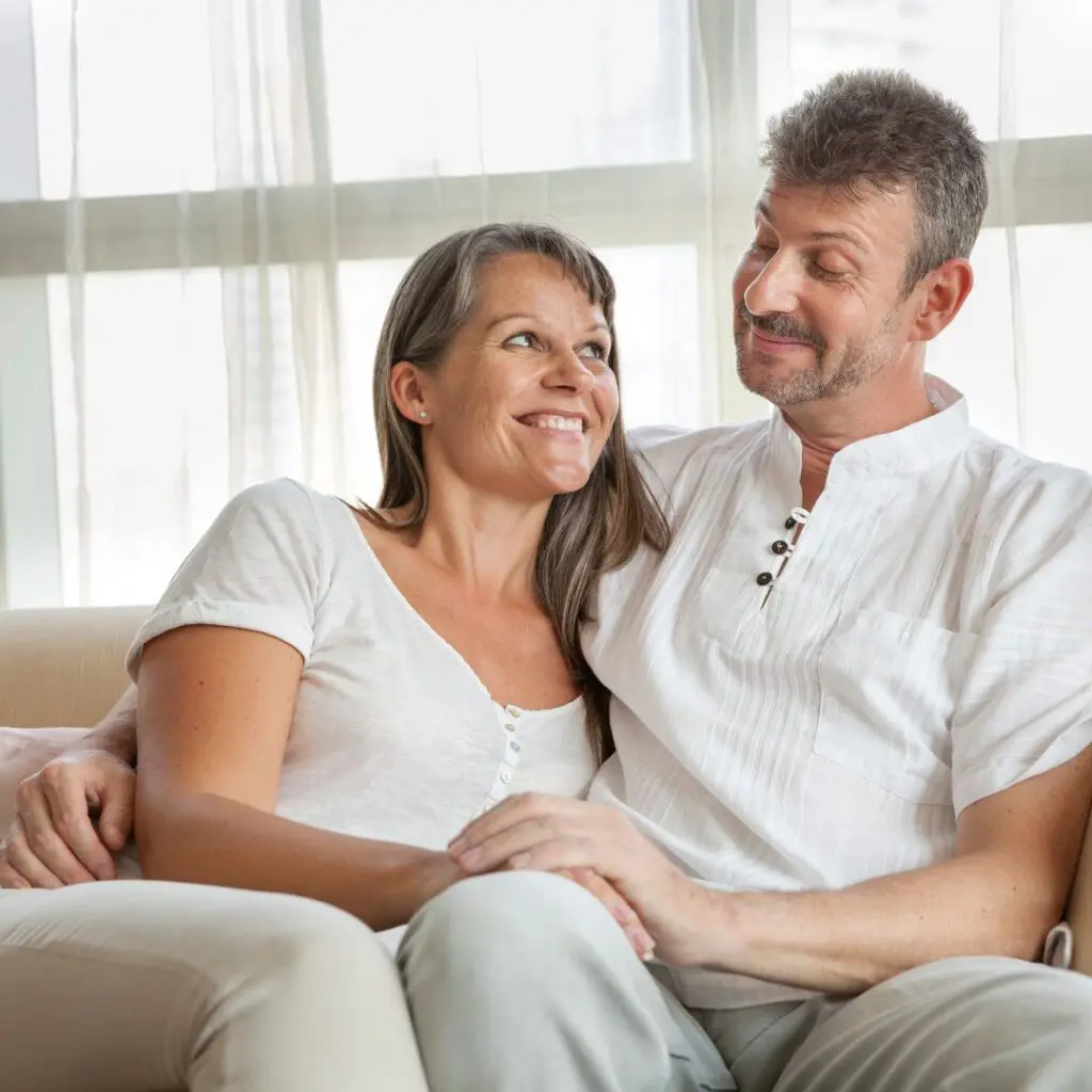 A middle-aged couple sit close together on a sofa, smiling and looking at each other affectionately. They both wear casual white clothes and appear relaxed and happy in a bright room with large windows.