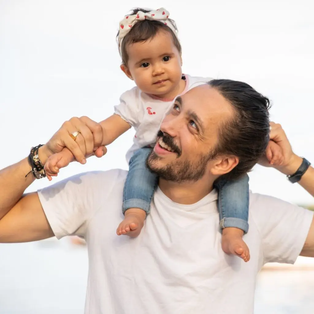 A man with a beard smiles as he carries a baby girl on his shoulders. The baby, wearing a white shirt, blue jeans, and a bow headband, holds his hands. Both are outdoors with a bright, blurred background.