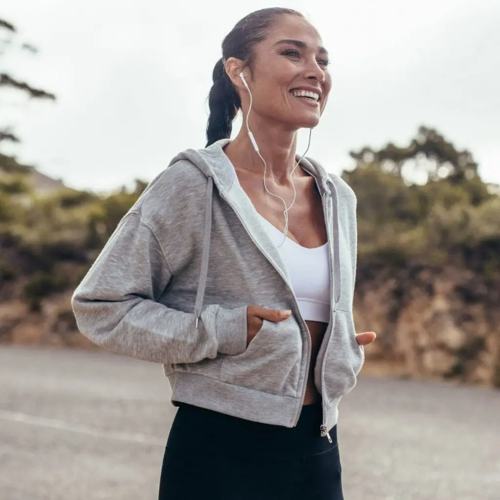 Woman smiling while jogging outdoors after Laser Eye Surgery, wearing a grey hoodie, white sports bra, and black leggings with earphones in. Trees and a cloudy sky form the background after laser eye surgery at centre for sight