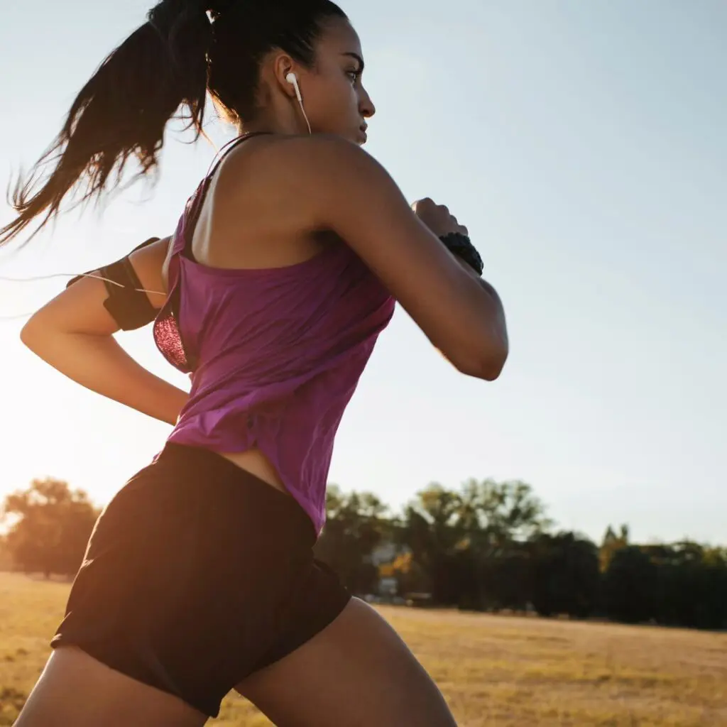 A young woman, post-LASIK, runs outdoors in a park at sunset, wearing a purple vest, black shorts, and earphones, her hair in a ponytail. Trees and grass frame the scene as she enjoys clear vision on her run.