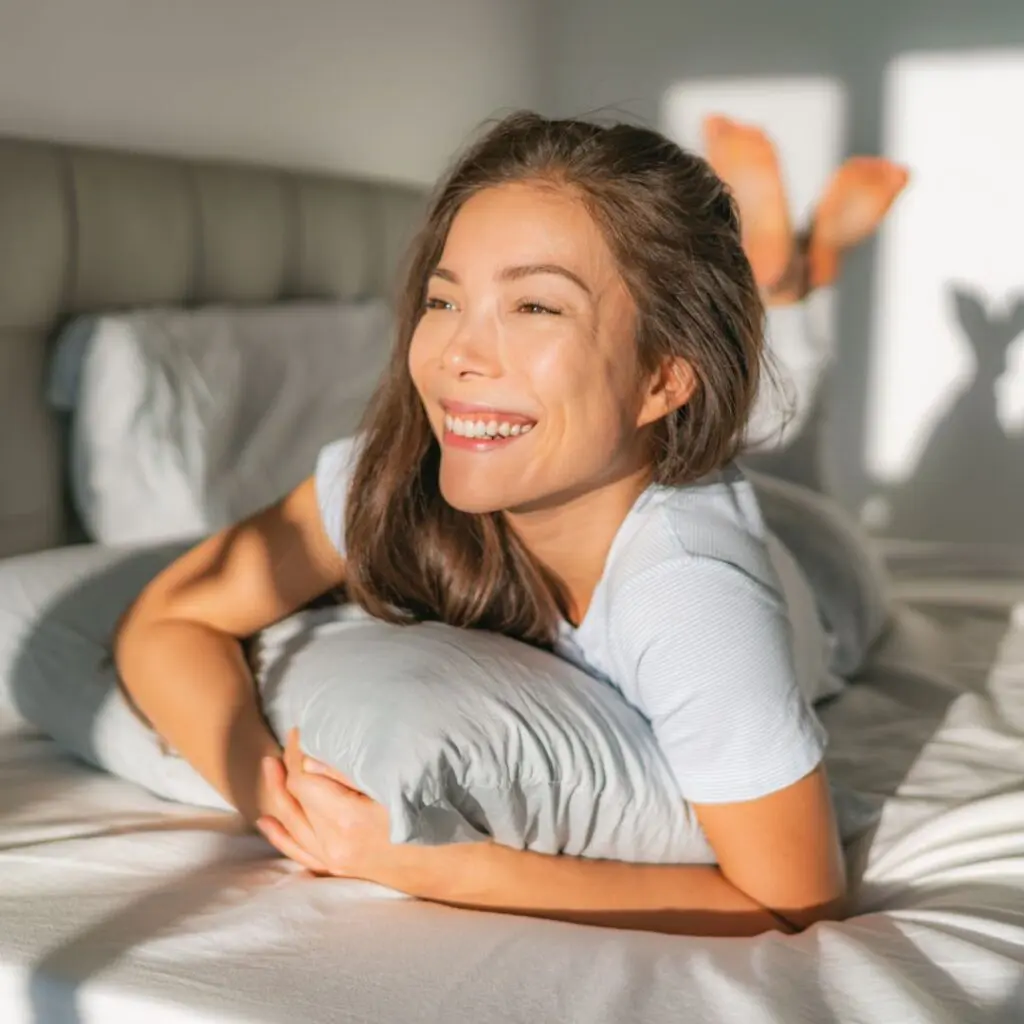A woman with long brown hair is lying on her stomach on a bed, hugging a pillow and smiling brightly—her clear vision after LASIK letting her enjoy the sunlight streaming through the window and casting soft shadows on the wall behind her.
