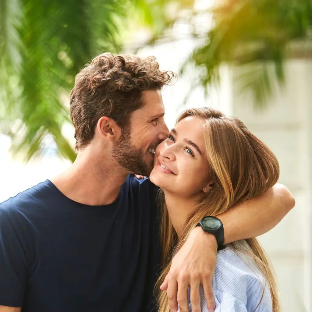 A man with curly hair embraces a smiling woman outdoors, both looking happy. The man wears a navy shirt and watch, whilst the woman, possibly enjoying her clear post-LASIK vision, gazes upward. Green foliage is blurred in the background.