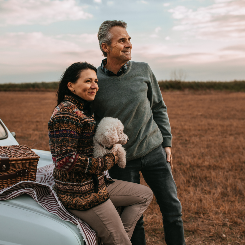 Man and woman couple in cut hay field standing looking at the view with dog in arms leaning against car with hamper for picnic