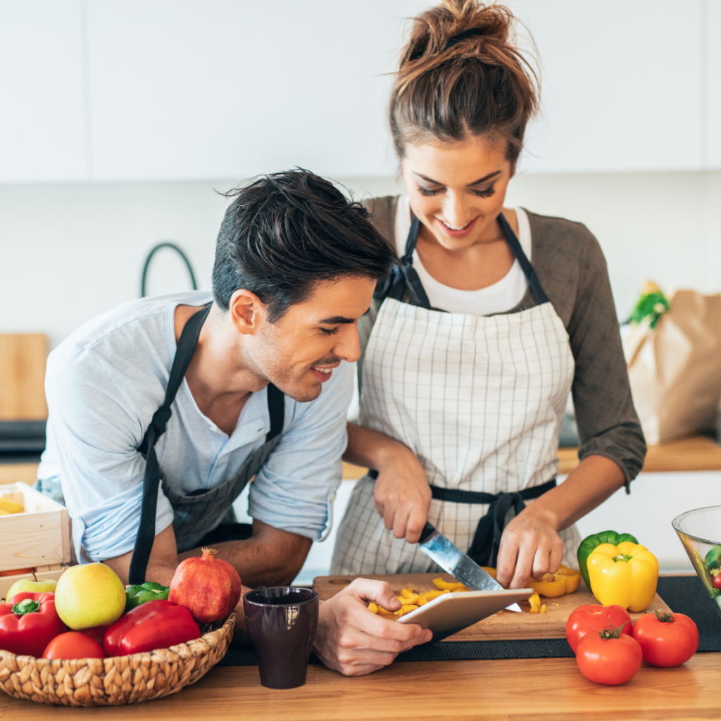 Couple cooking with fruit and vegetables in the kitchen with aprons on smiling following a recipe