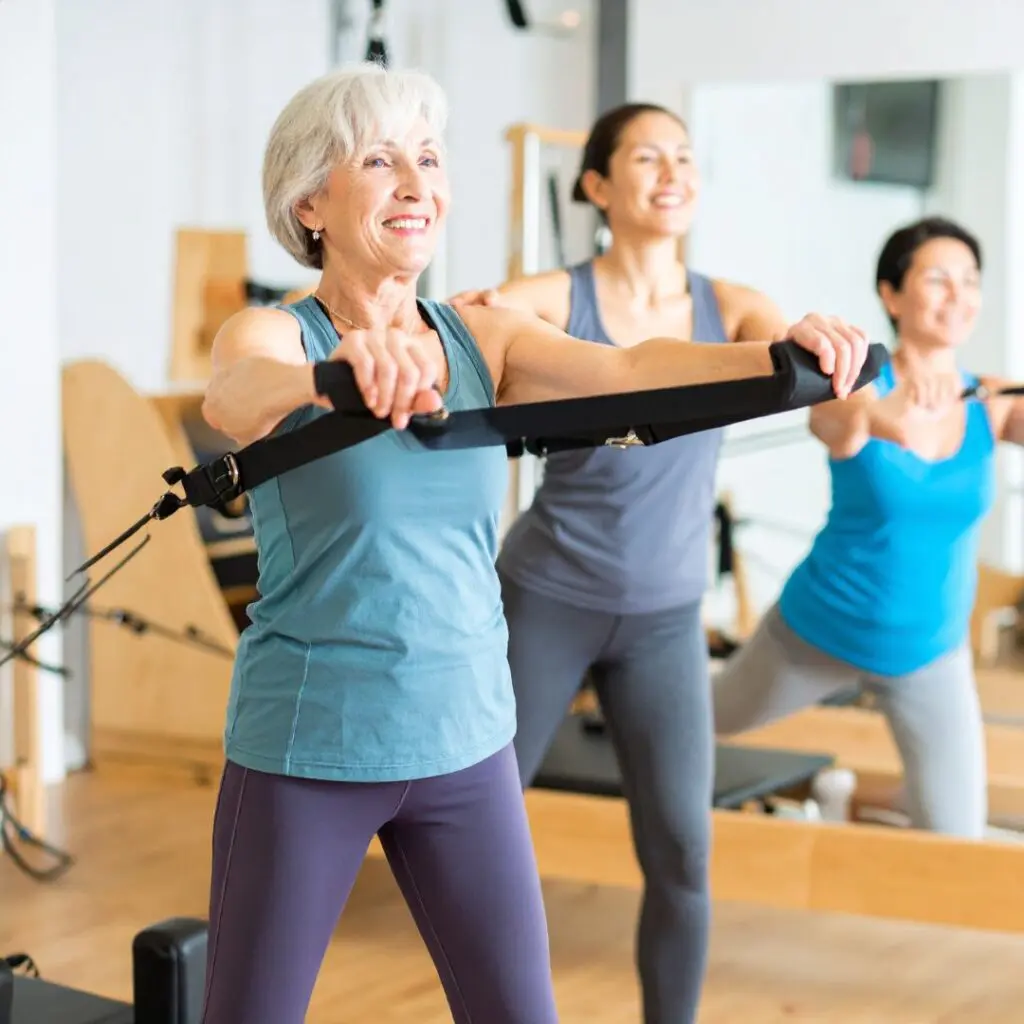 Three women in sportswear are exercising on Pilates reformer machines in a bright studio. The woman at the front, who previously underwent corneal transplantation at Centre for Sight, smiles as she stretches with resistance bands; two women are visible behind her.