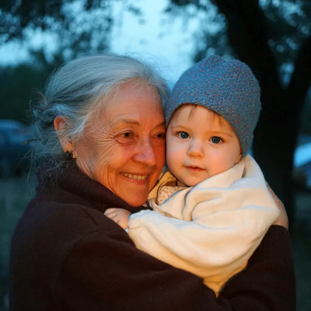 An elderly woman with grey hair, who recently underwent corneal transplantation at Centre for Sight, hugs a baby in a blue knitted hat and white blanket. Both are smiling outdoors in soft evening light with trees in the background.