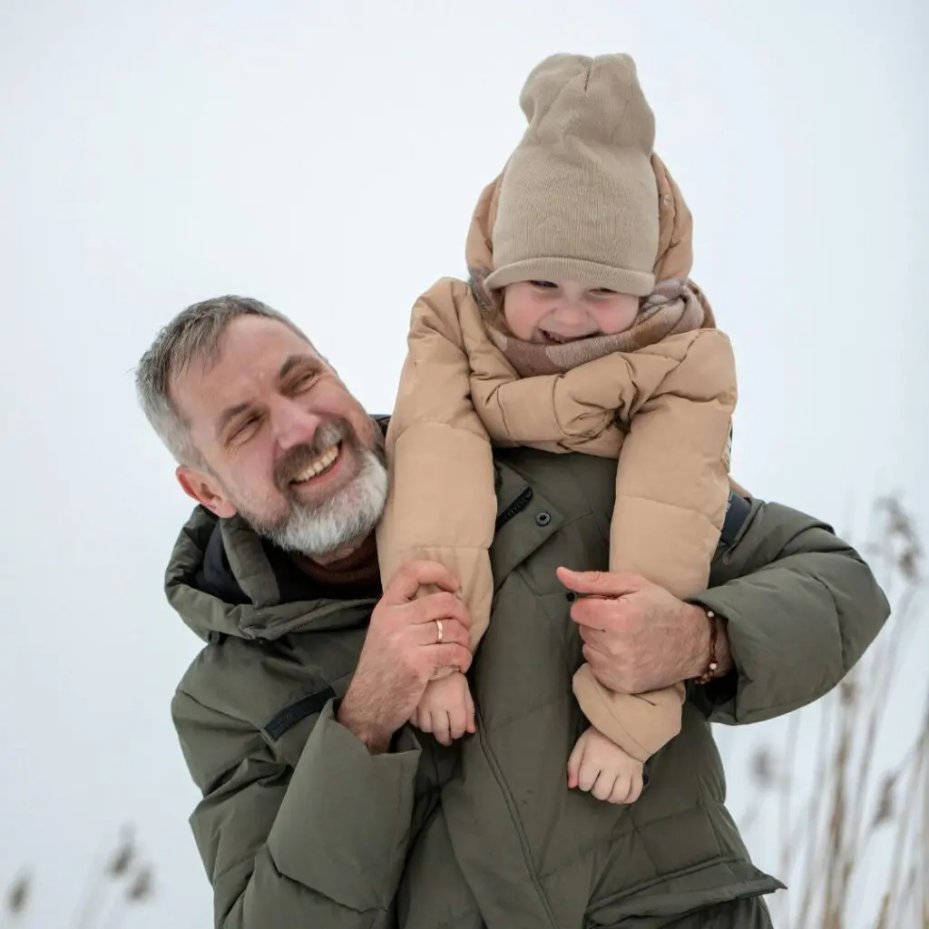 A smiling man with grey hair and beard, restored by corneal transplantation at Centre for Sight, carries a happy child in a beige winter coat and hat on his shoulders outdoors on a snowy day.