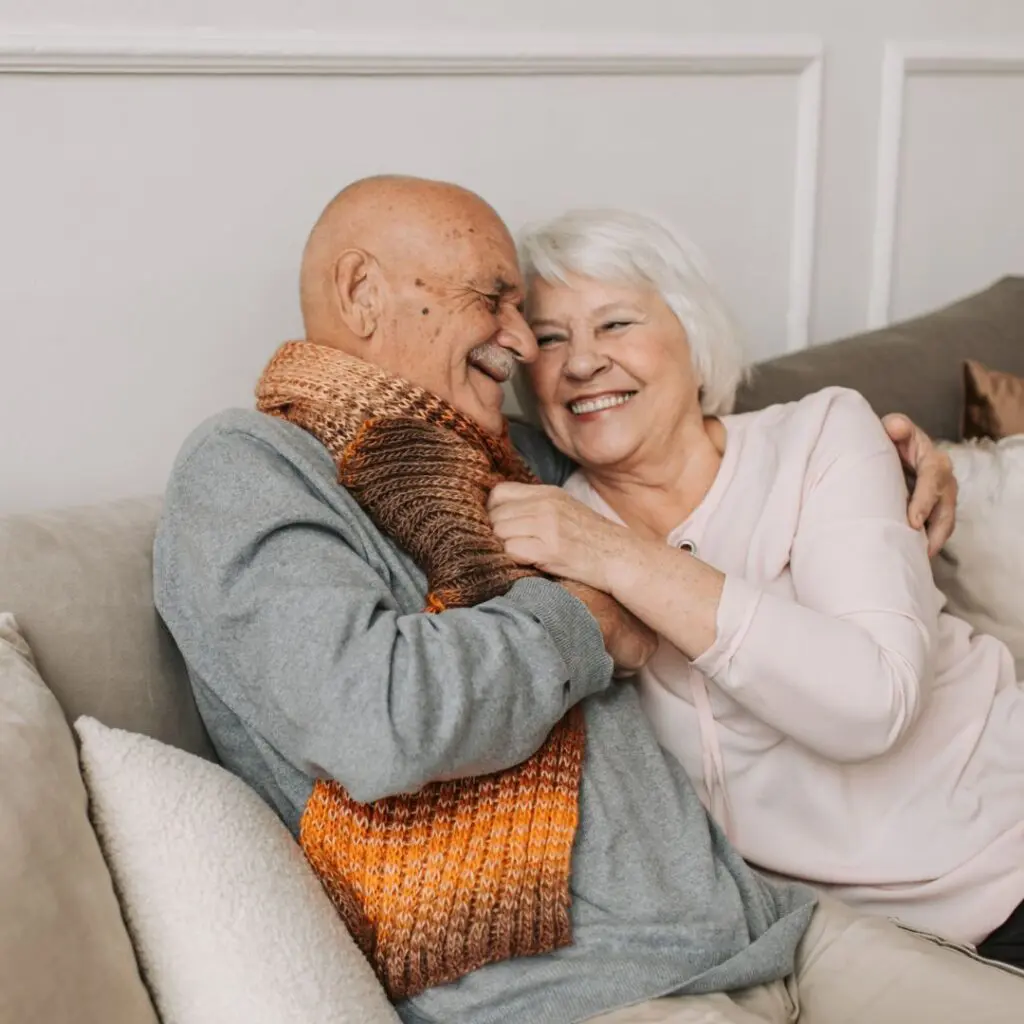 An elderly couple sits on a sofa, embracing and smiling warmly at each other, their happiness shining through after the man's successful corneal transplant at Centre for Sight. He wears a grey jumper and brown-orange scarf; she, a light pink top.