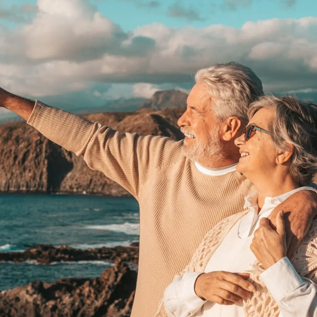 A smiling older couple stands by the sea, with rocky cliffs in the background. The man, who recently recovered from corneal transplants at centre for sight, points towards the horizon while embracing the woman. Both appear happy and relaxed under a partly cloudy sky.
