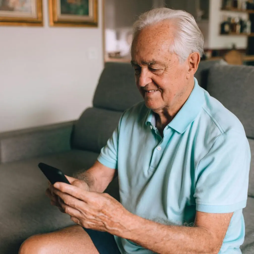 An elderly man with white hair is sitting on a sofa, smiling whilst looking at a mobile phone in his hands. Having recently undergone corneal transplants at Centre for Sight, he appears relaxed and content in a cosy living room, wearing a light blue polo shirt.