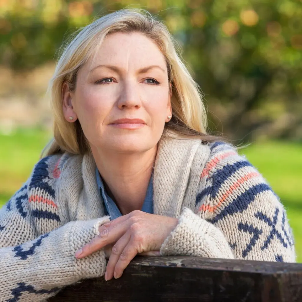 A middle-aged woman with blonde hair, wearing a patterned knitted jumper, leans on a wooden surface outdoors, looking thoughtfully into the distance, perhaps reflecting on her experience with corneal transplants at Centre for Sight. The background is blurred greenery.