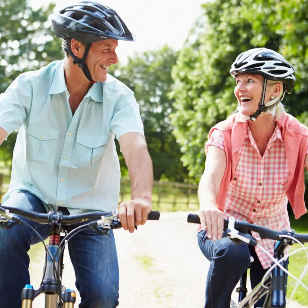 Two older adults wearing helmets ride bicycles on a sunny day, smiling at each other—enjoying the outdoors thanks to renewed vision after corneal transplants at centre for sight, with green trees and grass in the background.