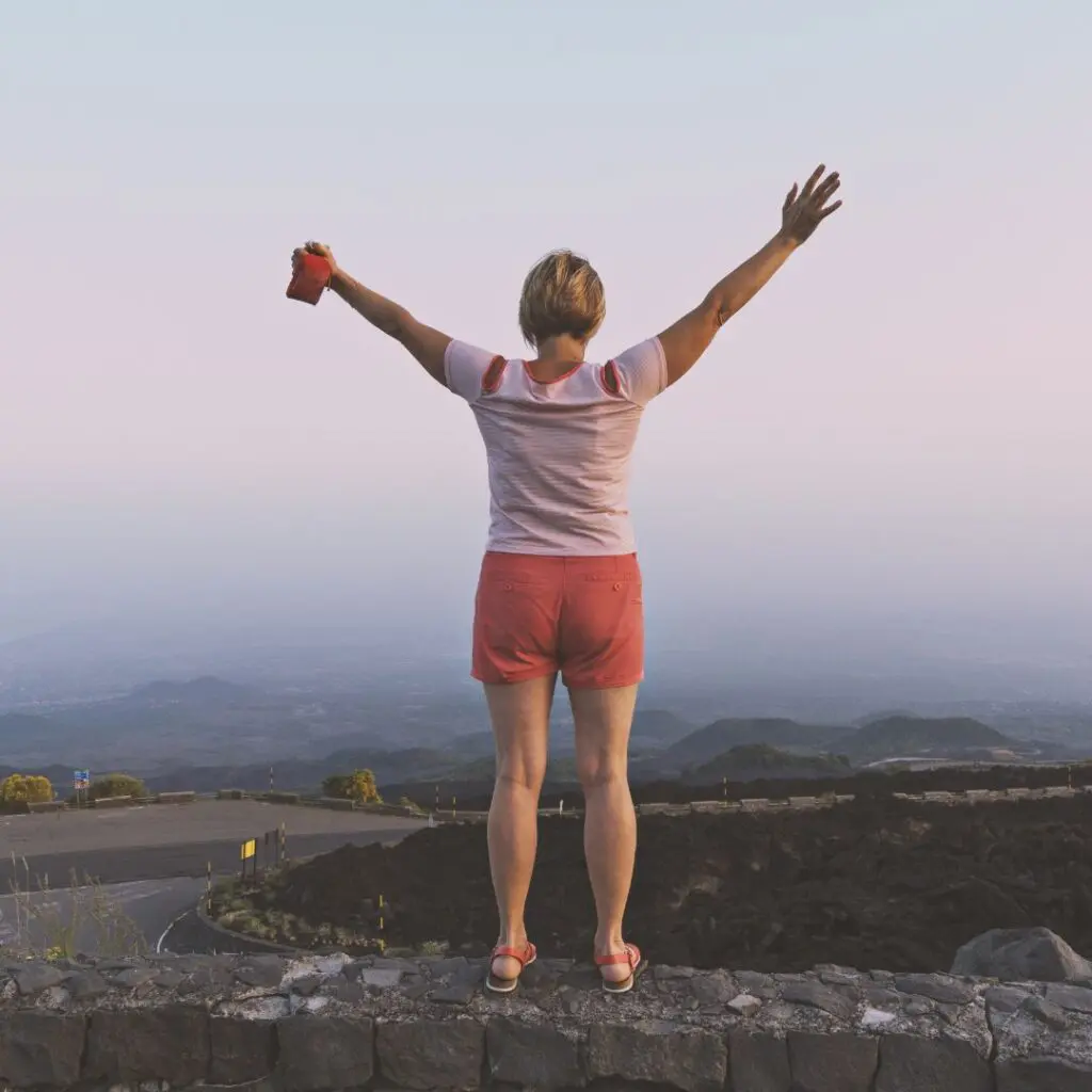 A person stands on a stone wall with arms raised, facing a scenic landscape of mountains and a winding road at sunset. They wear a pink shirt and shorts, holding a red object in one hand.