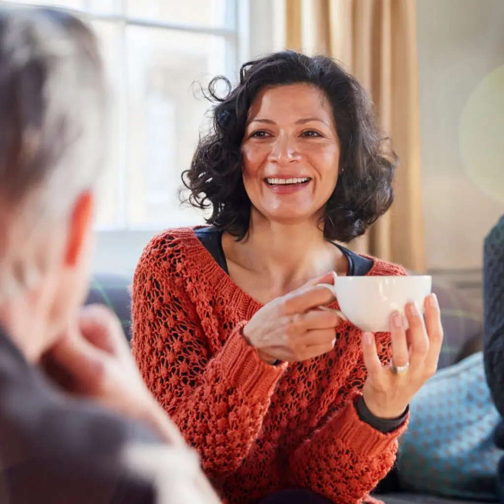 A woman with curly dark hair, wearing an orange jumper, smiles while holding a white mug. She is sitting indoors and appears to be having a friendly conversation with someone in the foreground.