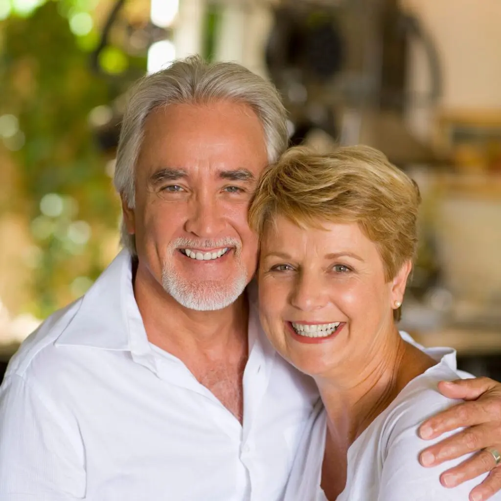 An older couple with fair hair and bright smiles embrace each other, both wearing white shirts, standing indoors with a blurred background of greenery and soft lighting.