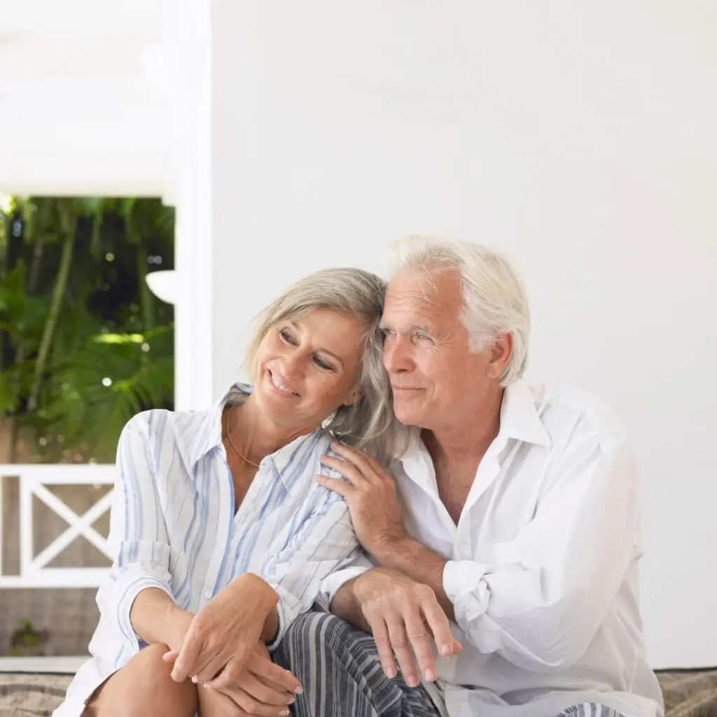An older couple sit closely together, smiling and relaxed, with the woman resting her head on the man's shoulder. After DSAEK surgery, they are dressed casually and appear happy in a bright, airy setting with greenery outside. cataract eye surgery