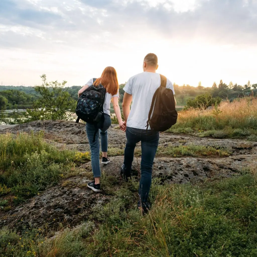 A couple with rucksacks holds hands while hiking on a rocky path surrounded by grass and bushes, enjoying the sunset—an adventure made clearer thanks to implantable contact lens technology.
