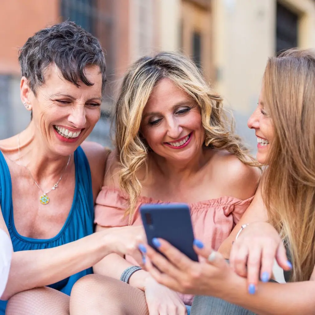 Three women sit close together outdoors, smiling and laughing as they look at a mobile phone—one sharing her recent DSAEK journey—enjoying a moment of friendship and connection.