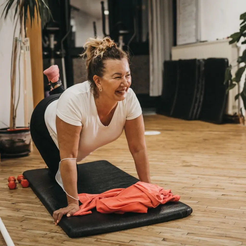 A woman smiles whilst exercising on a yoga mat indoors after her DSAEK procedure, wearing a white shirt and black leggings. An orange shirt and small weights are on the floor beside her, surrounded by wooden floors and fitness equipment.