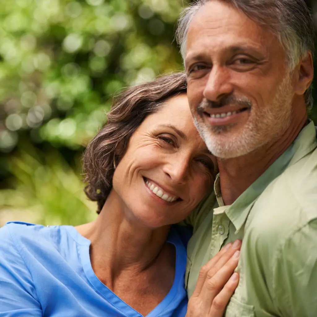 A smiling middle-aged couple embraces outdoors, with the woman resting her head on the man's shoulder. Both appear happy and relaxed after the woman's successful DSAEK surgery, standing in front of a blurred green background.