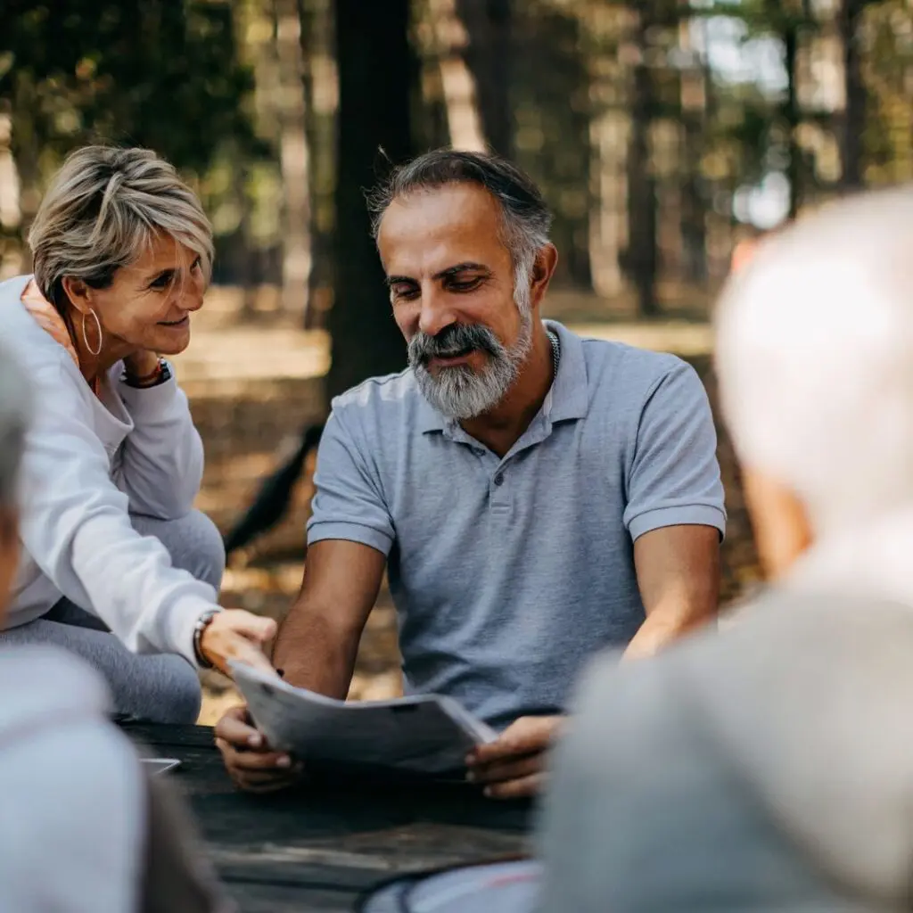 Two older adults sit at an outdoor table in a park, smiling and looking at a newspaper together, perhaps reading about advances like Femtosecond Laser DALK at Centre for Sight. Trees and sunlight fill the background, with two more people blurred in the foreground.