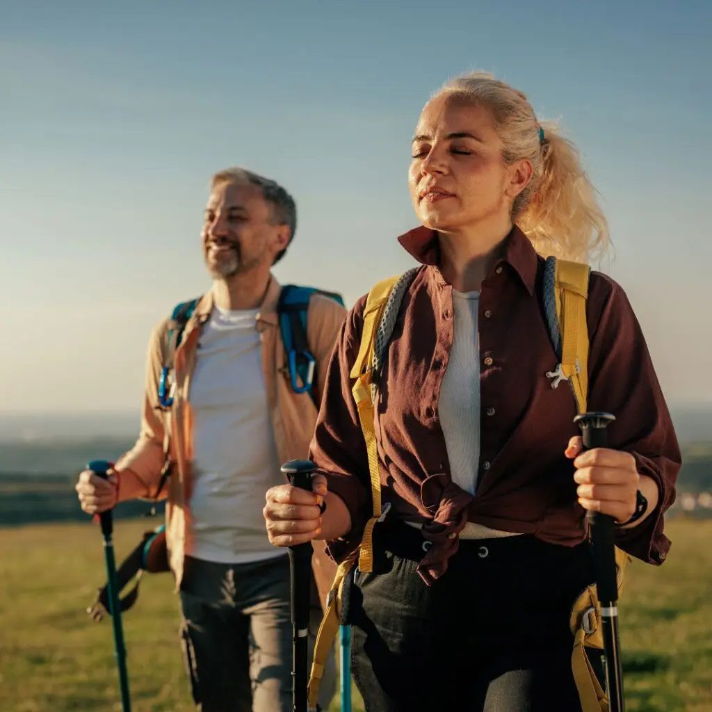 A woman and a man are hiking outdoors in sunny weather, using trekking poles and wearing rucksacks. The woman, refreshed after her Femtosecond Laser Dalk procedure at Centre for Sight, is in the foreground with her eyes closed; the man smiles in the background.