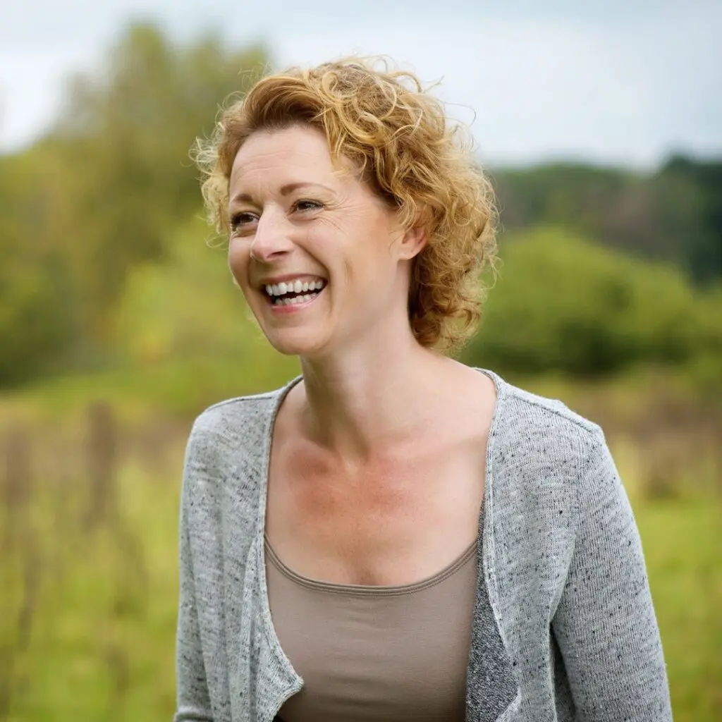 A woman with short, curly blonde hair smiles and laughs outdoors, wearing a light grey cardigan over a tan top. She stands in front of a blurred green field and trees, radiating joy—much like the positive outcomes seen after Femtosecond Laser Dalk.