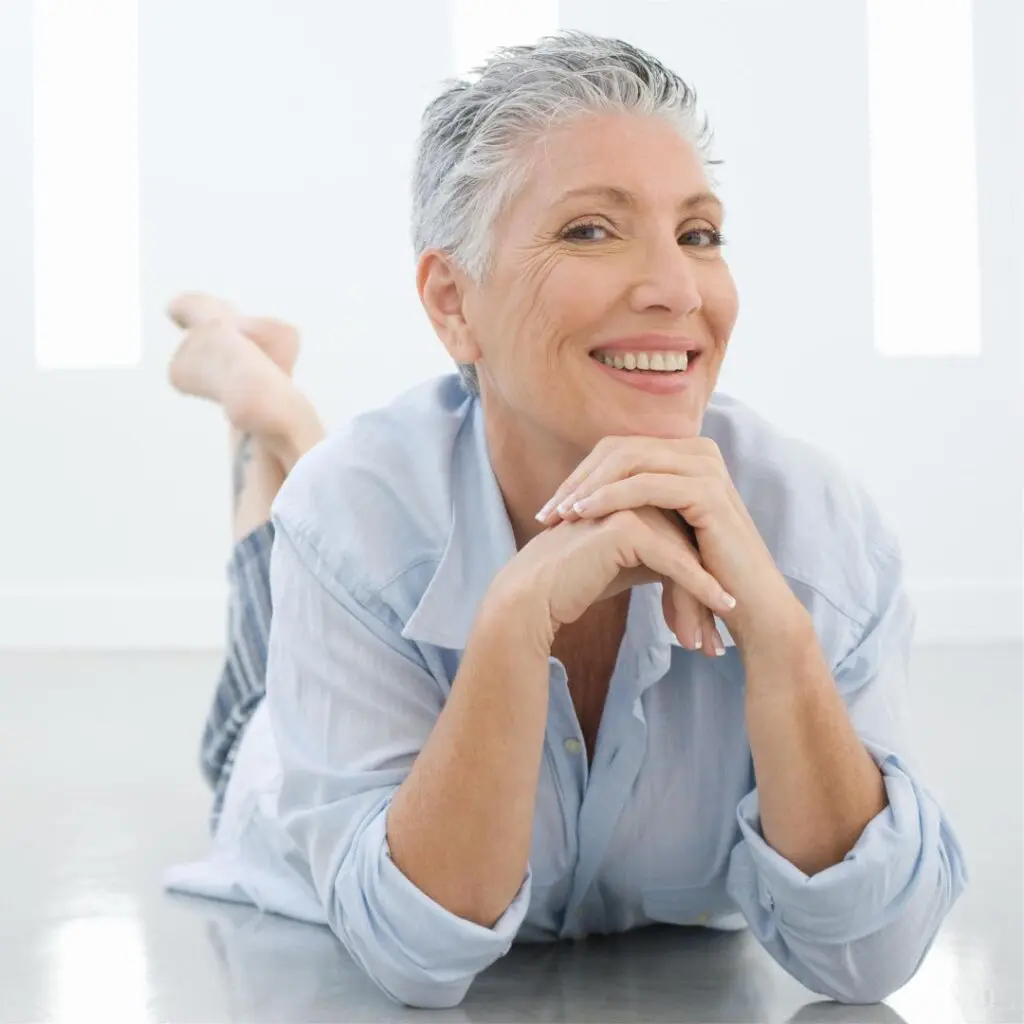 An older woman with short grey hair lies on her stomach on a shiny floor, smiling warmly at the camera. She wears a light blue shirt and rests her chin on her hands in a bright, modern room after a successful Femtosecond Laser Dalk procedure at Centre for Sight.