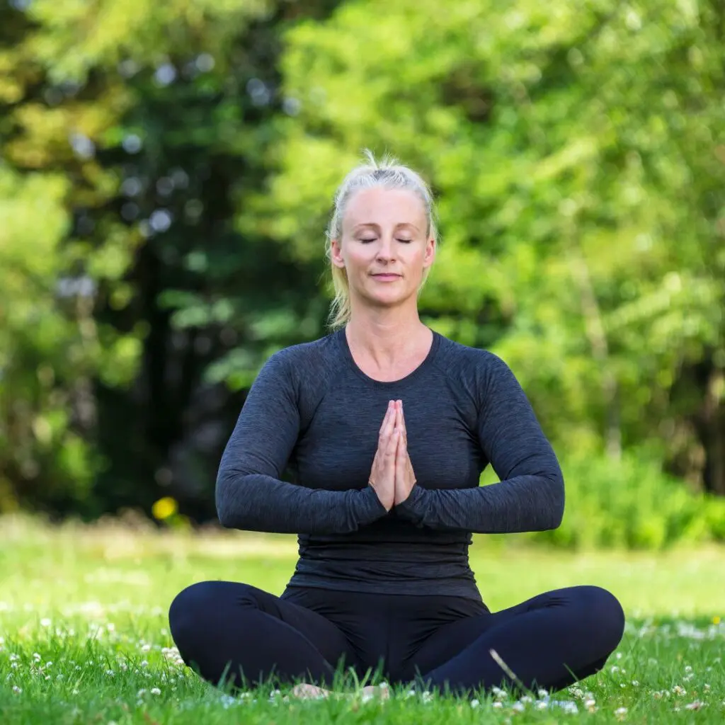 A woman sits cross-legged on grass outdoors, eyes closed and hands pressed together in a prayer pose. She wears a black long-sleeve top and leggings, surrounded by greenery and trees in the background.