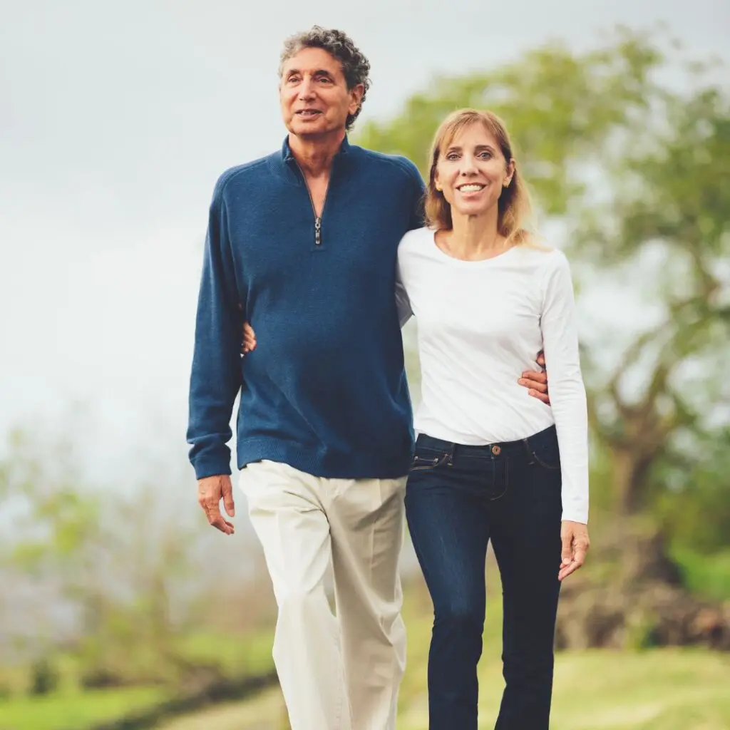 A middle-aged couple walks outdoors, smiling and arm in arm. The man wears a blue jumper and light trousers; the woman wears a white long-sleeve top and dark jeans. Green trees and grass are in the background.