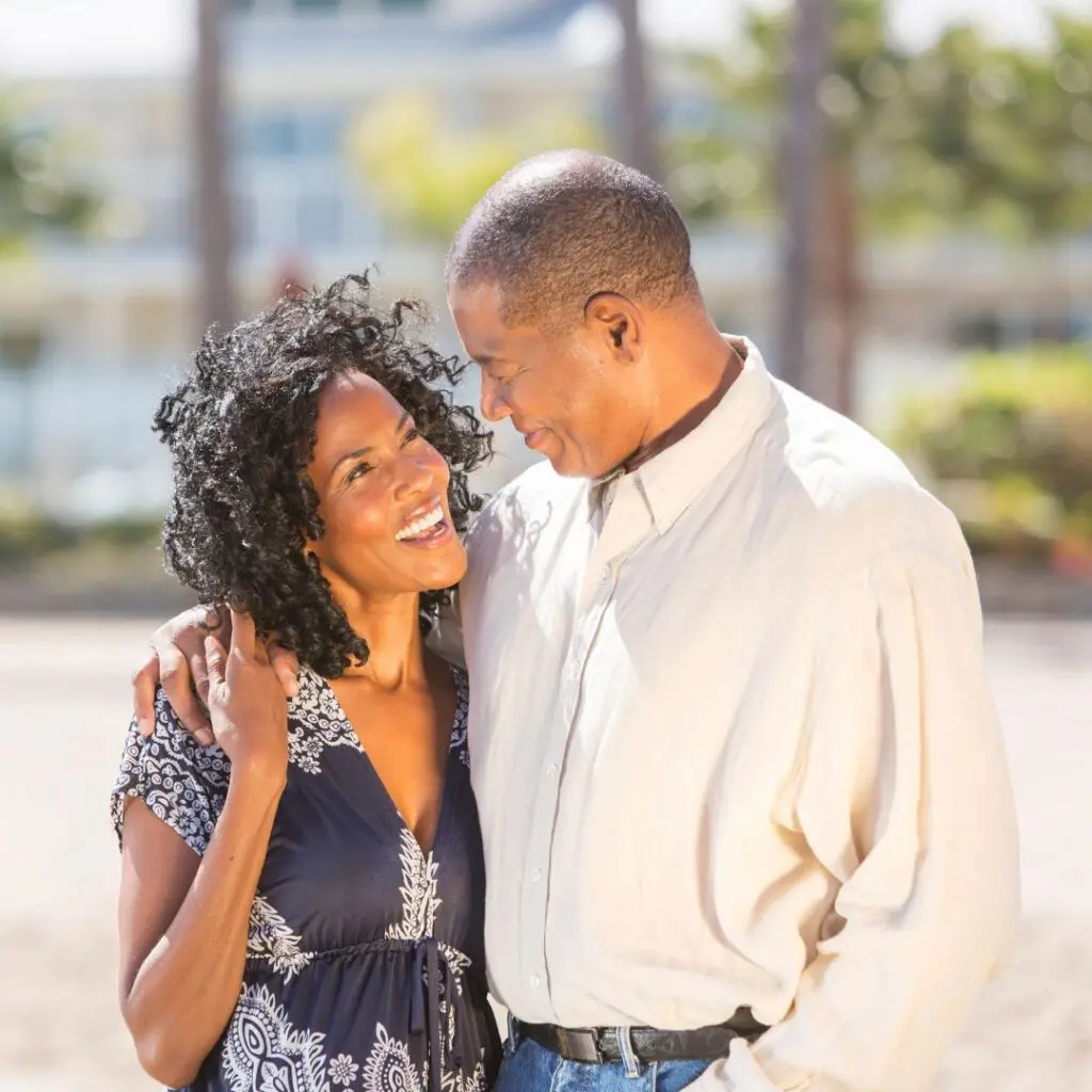 A smiling couple stand close together outdoors, with the woman’s arm round the man’s shoulder. They look at each other affectionately, with blurred greenery and buildings in the background.