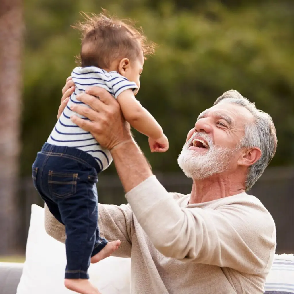 An older man with grey hair and a beard, refreshed after cataract eye surgery, smiles joyfully as he lifts a laughing baby in the air outdoors. The baby wears a striped shirt and jeans, with greenery blurred in the background.