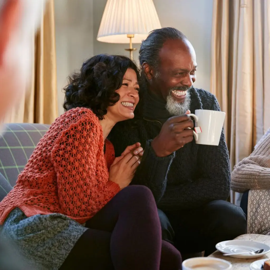 A smiling couple sit close together on a sofa, sharing a laugh while holding mugs after Femtosecond Laser PK at Centre for Sight. They appear relaxed and happy, with a lamp and curtains in the cosy background.