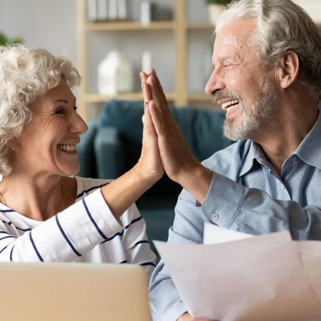 An older man and woman smile and high-five each other while sitting indoors, holding papers, with a laptop in front of them, celebrating the success of cataract eye surgery. They appear happy and triumphant.