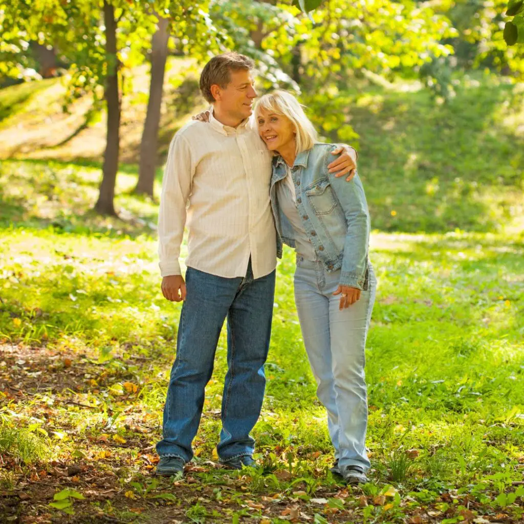An older couple stands close together in a sunlit park, smiling and embracing after pk surgery. They are dressed casually in jeans and light jackets, surrounded by green grass and trees.