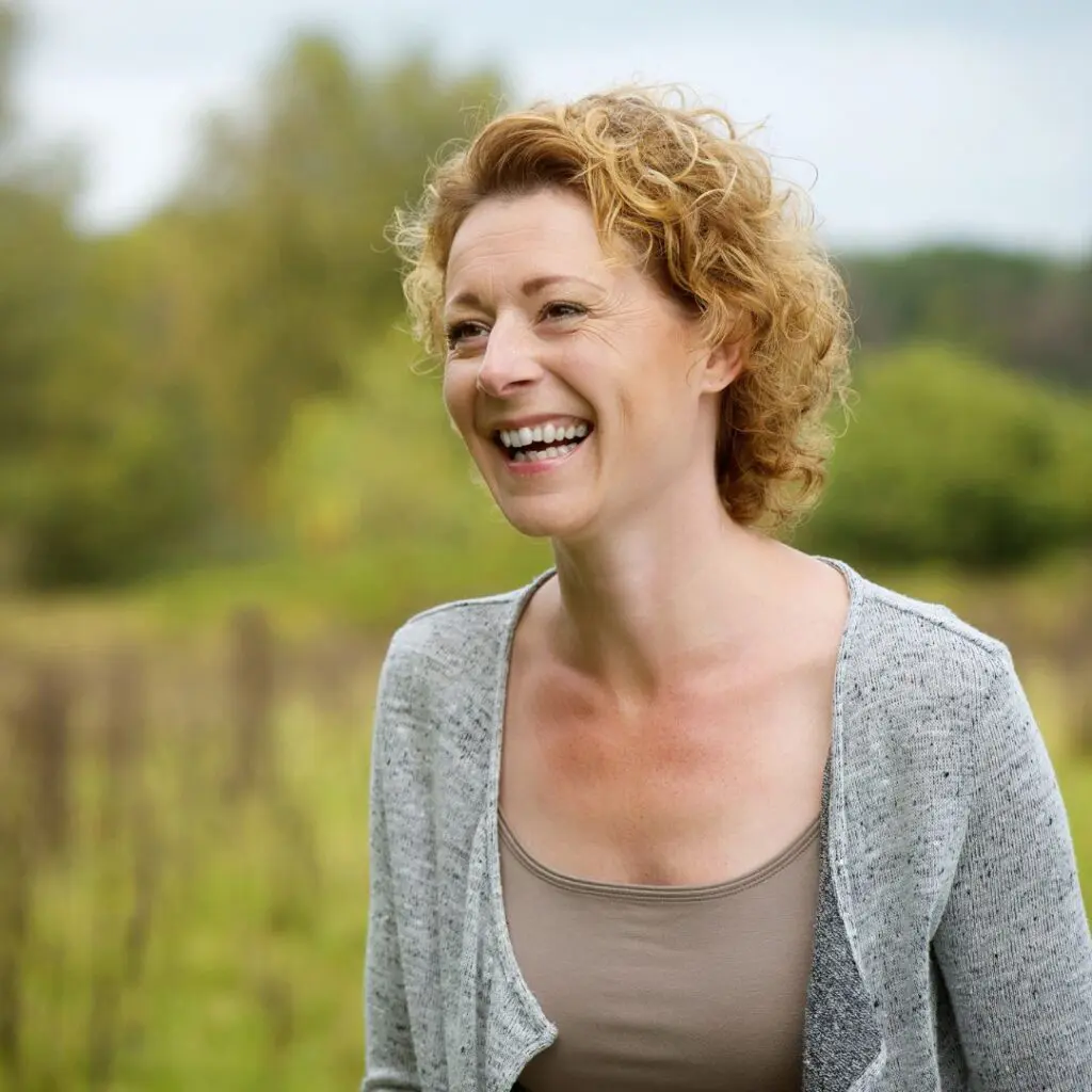 A woman with curly blonde hair smiles whilst standing outdoors in a field, wearing a beige top and a light grey CAIRS cardigan. Green trees and a cloudy sky are visible in the background.