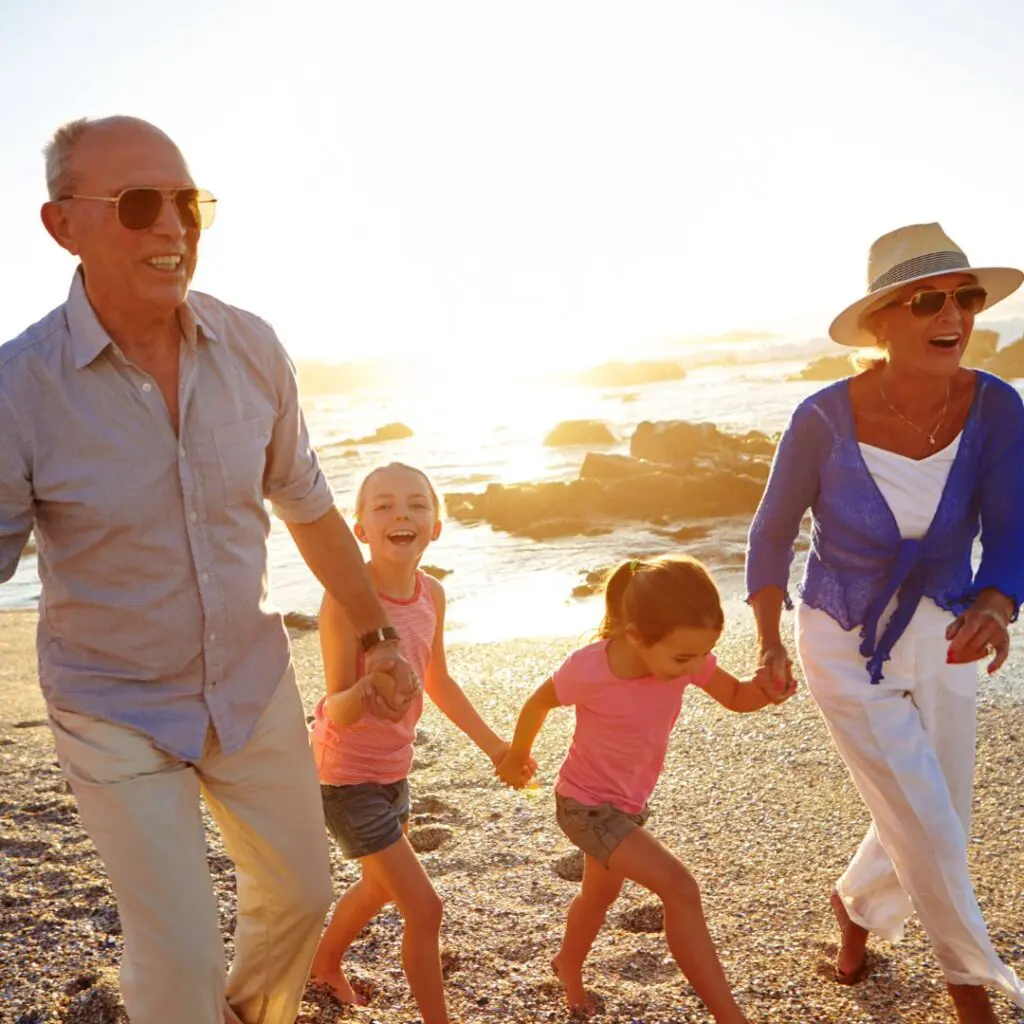 Two adults and two children smile and hold hands while walking along a sunny, sandy beach with rocks in the background. The adults wear sunglasses and hats, enjoying a cheerful day together exploring CAIRS coastal beauty.