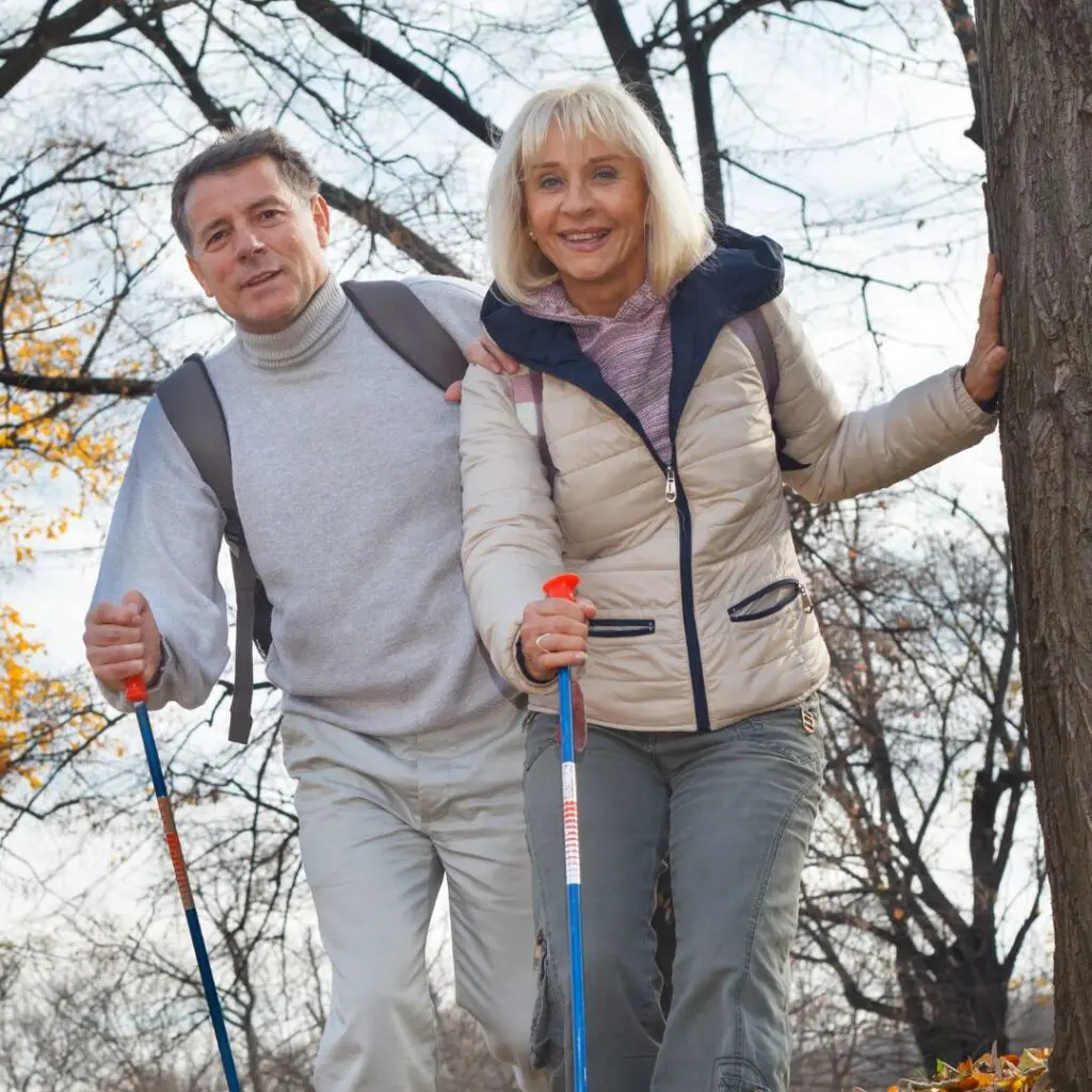 An older man and woman, both smiling, are hiking outdoors with rucksacks and walking sticks. Dressed warmly in jackets, they enjoy the crisp air among bare trees on a cool autumn day—part of their healthy CAIRS lifestyle.