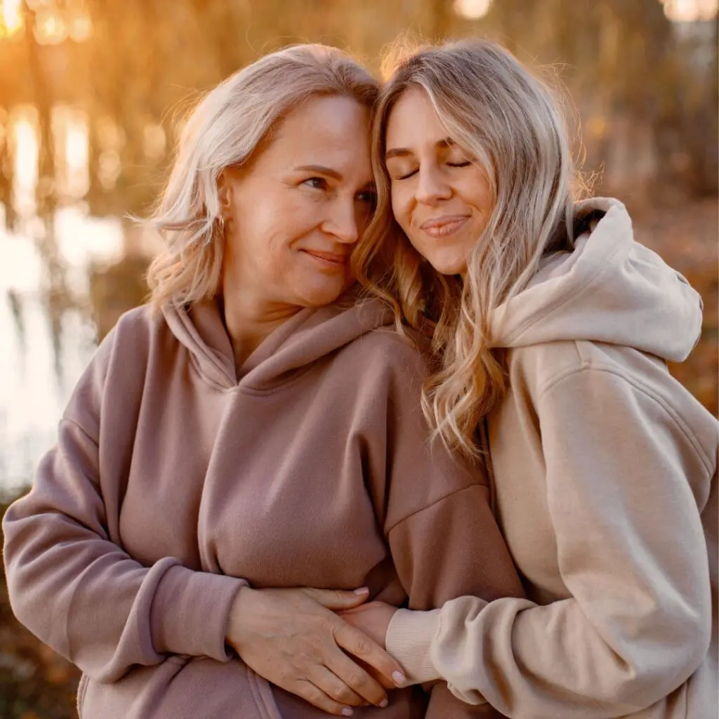 Two women in hoodies embrace outdoors at sunset. One smiles softly at the other, who leans in with eyes closed, both appearing happy and content—sharing a peaceful moment as warm light glows through trees, like collagen cross-linking for the soul.