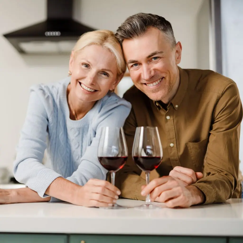 A smiling couple sit close together at a kitchen worktop, each holding a glass of red wine. They lean in, looking happy and relaxed in a modern kitchen—a perfect moment to toast to love and the benefits of collagen cross-linking.
