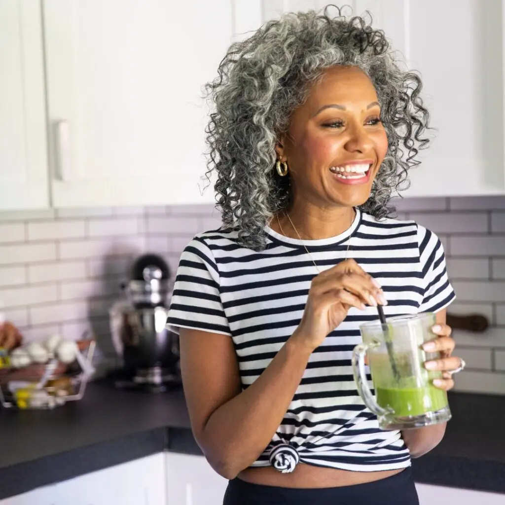 A smiling woman with curly grey hair holds a glass of green smoothie in a modern kitchen, wearing a black and white striped shirt. Kitchen appliances and eggs are visible on the counter, hinting at a collagen crosslinking-friendly lifestyle.