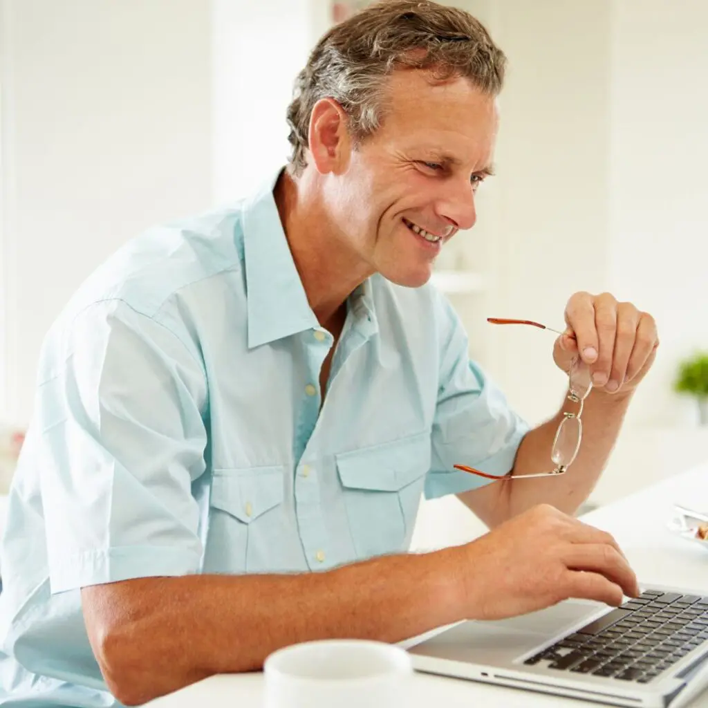 A middle-aged man in a light blue shirt smiles whilst using a laptop. Holding his glasses, he appears to be researching collagen cross-linking at his desk, with a white coffee mug in the foreground.