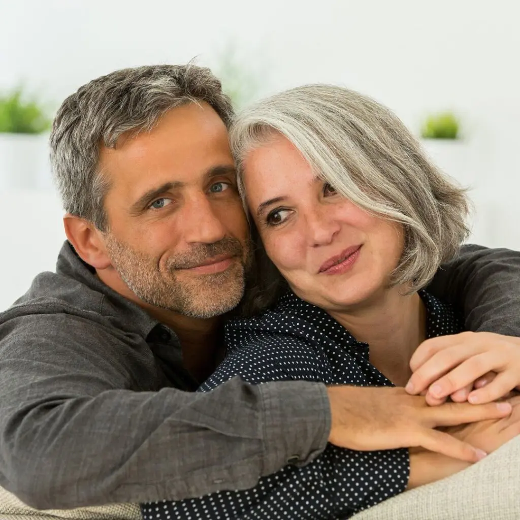 A middle-aged couple sit closely together on a sofa, smiling softly after penetrating keratoplasty surgery. The man has short dark hair with some grey, while the woman has chin-length grey hair. They look relaxed and content, with his arm around her shoulders.